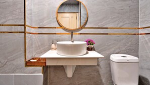 Une salle de bain avec un mur en marbre, un miroir rond, un lavabo sur pied, une poubelle et un petit panier en osier.