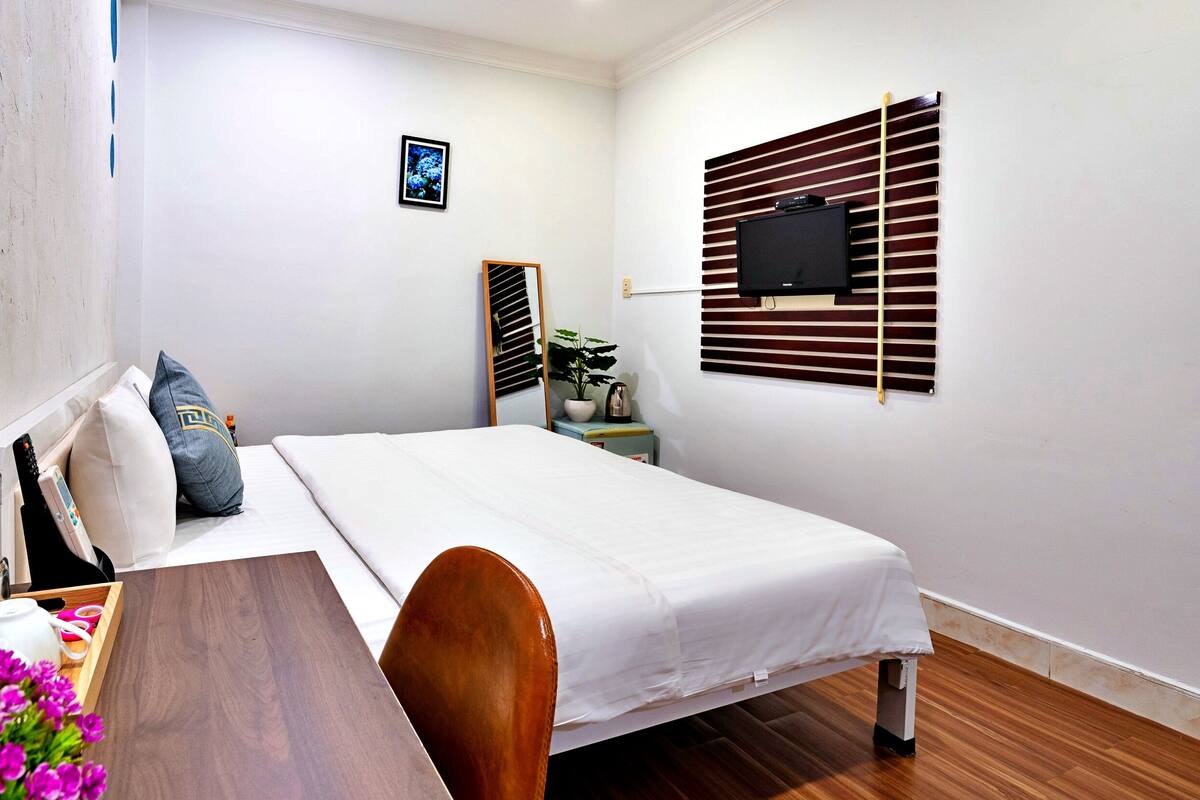 Ein ordentlich eingerichtetes Hotelzimmer mit Bett, Nachttisch, Spiegel, an der Wand montiertem Fernseher und einer kleinen Topfpflanze.