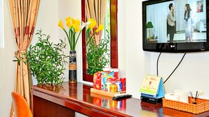 Ein Hotelrezeptionstresen mit Holzbeschlag, ein an der Wand befestigter Fernseher, ein Stuhl und Topfpflanzen.