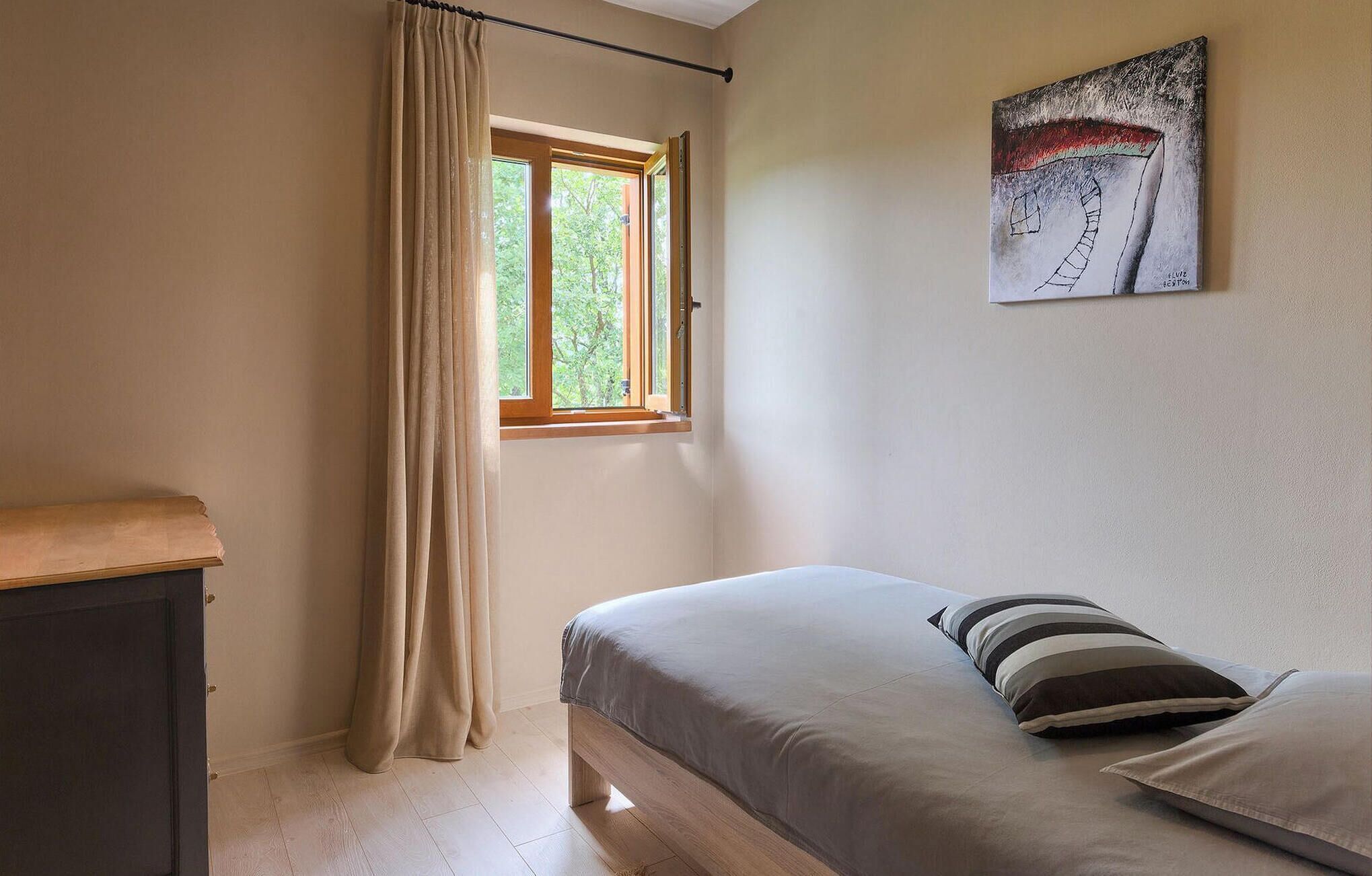Ein Schlafzimmer mit Bett, einem Fenster mit Vorhängen, einem Bild an der Wand und einem hölzernen Kopfteil.
