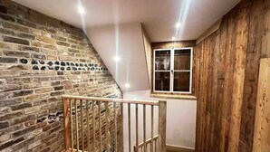 A loft bedroom with exposed brick walls, wooden paneling, a wooden railing, and a window with a view. - The Stables at Historic Grade ll Listed Farm (Broadstairs)