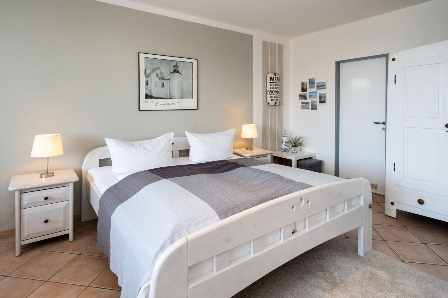 A bedroom with a bed, two bedside tables, a nightstand, a door, and a wall with a framed picture and a sign.