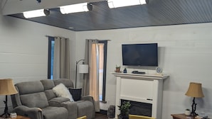 Ein Wohnzimmer mit einer Couch, einem Fernseher, einem Kamin, einem Esstisch mit Stühlen und einer Decke mit Lampen.