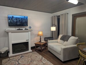 A living room with a fireplace, a television mounted above it, a sofa, a lamp, and a small table. - Cozy cottage located in rural Central Ohio. (Utica)