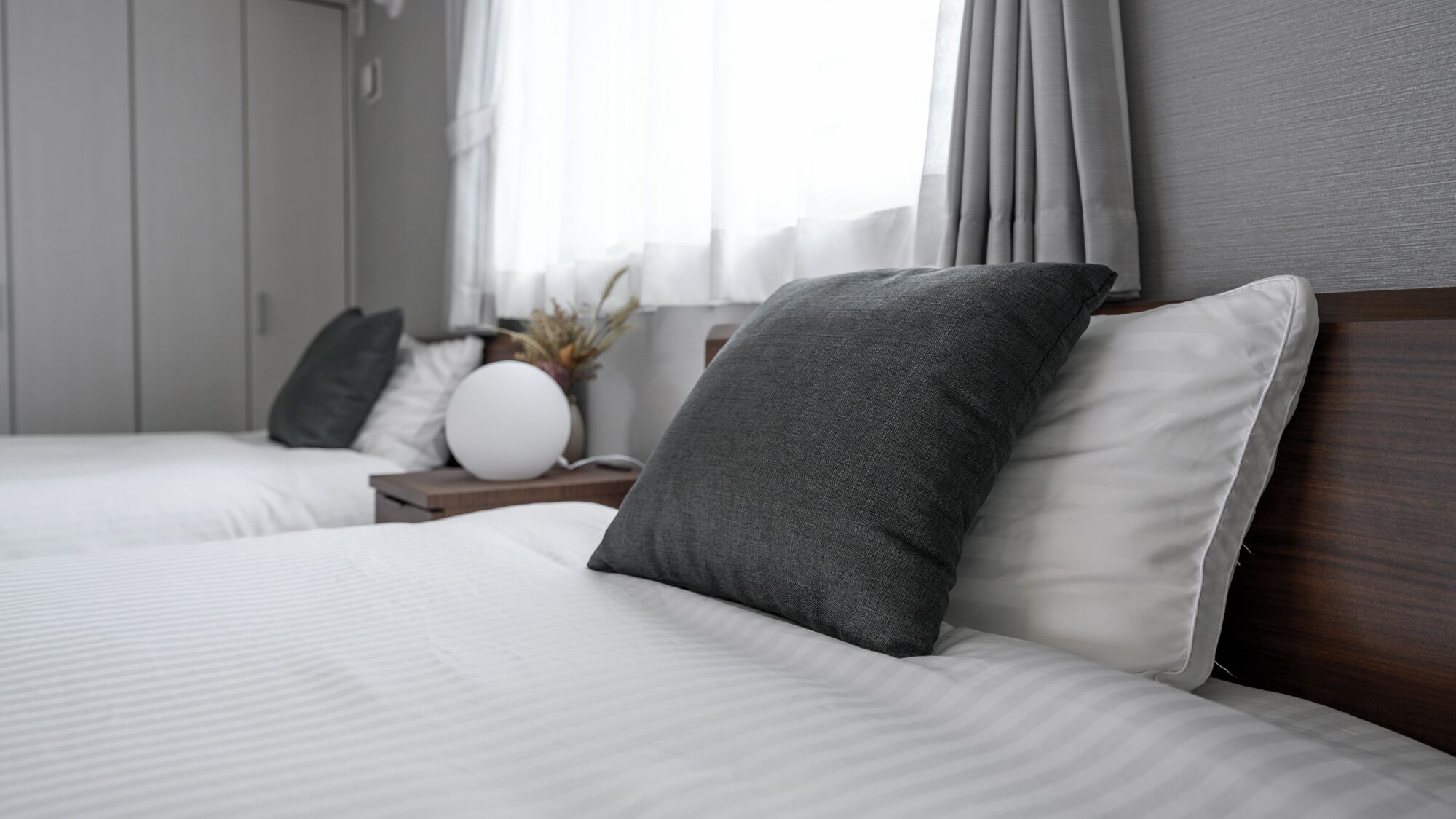 A hotel room with two beds, a nightstand, a white vase with dried plants, and a window with curtains.