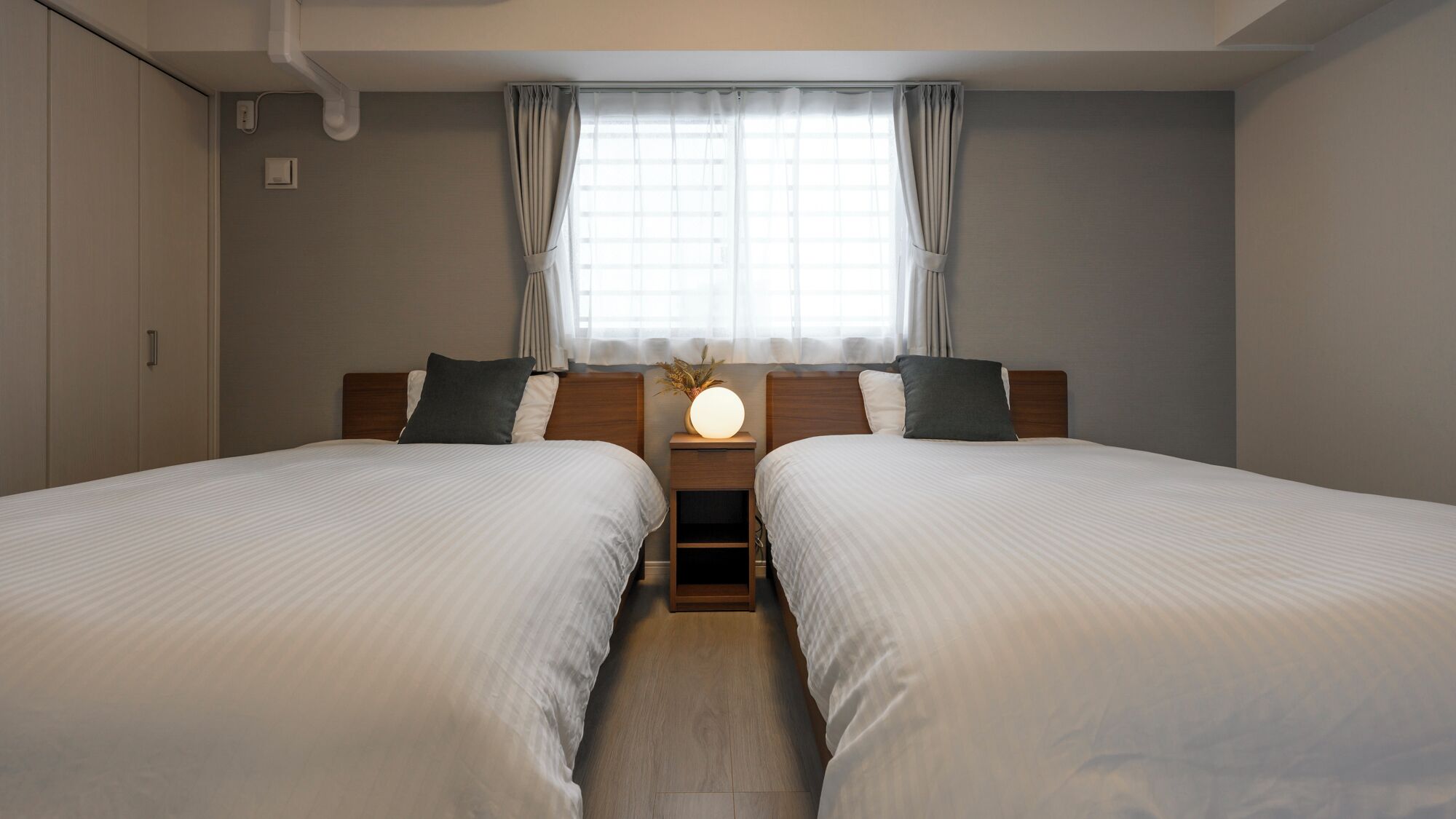 A hotel room with two beds, a bedside table with a lamp, a window with curtains, and a wall-mounted air conditioner.