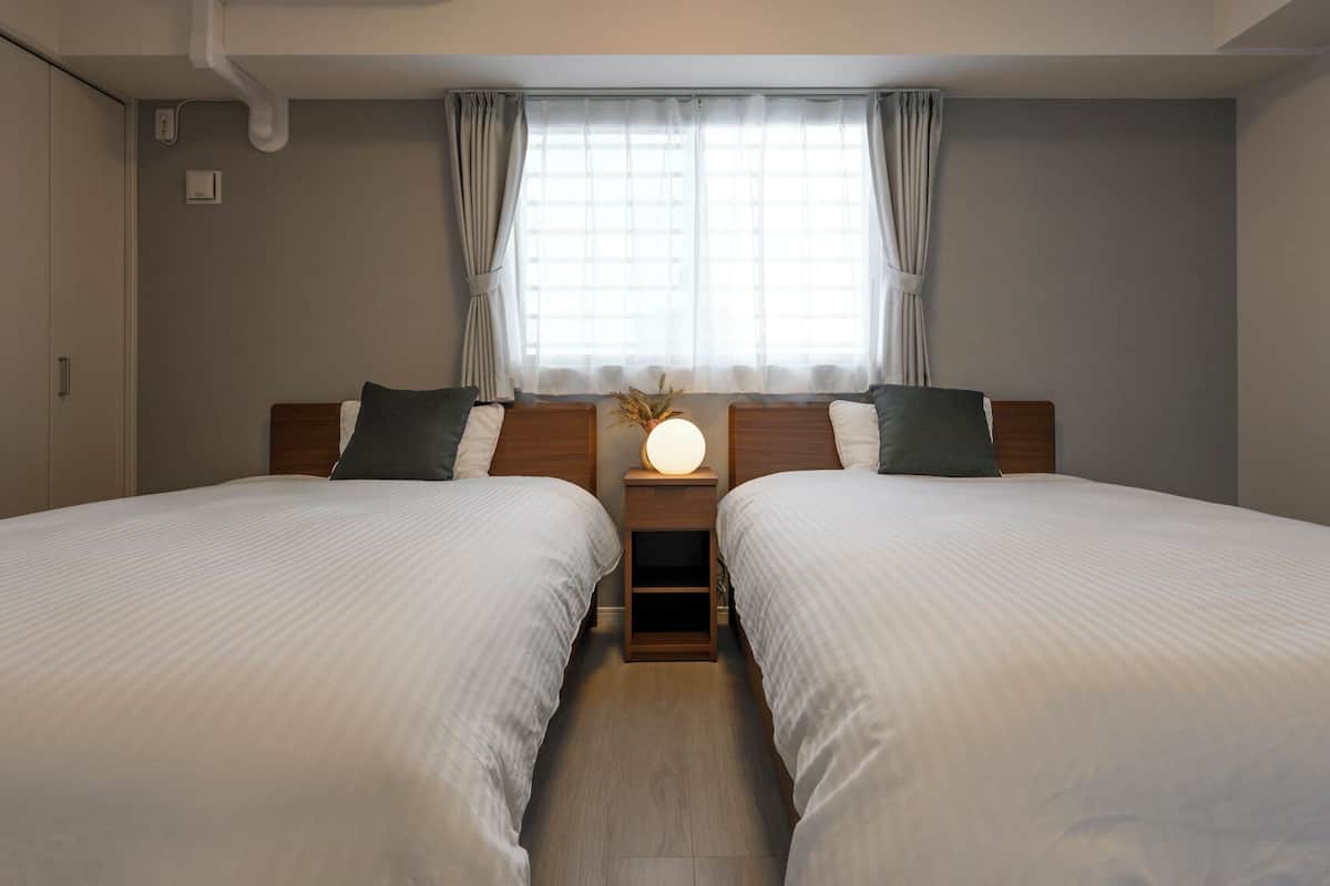A hotel room with two beds, a bedside table with a lamp, a window with curtains, and a wall-mounted air conditioner.