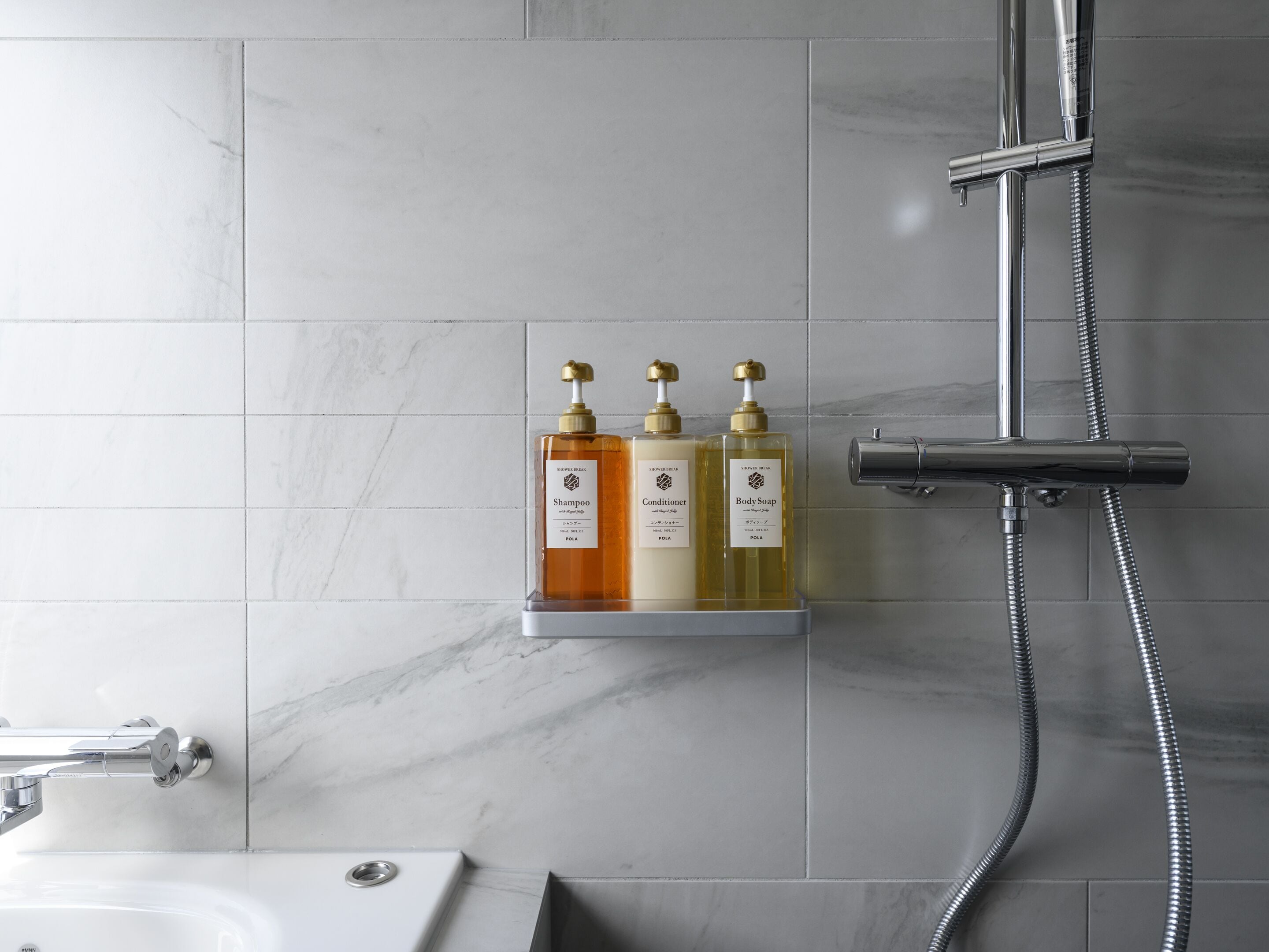 A bathroom with a marble floor, a shower with a handheld showerhead, and three bottles of toiletries on a shelf.