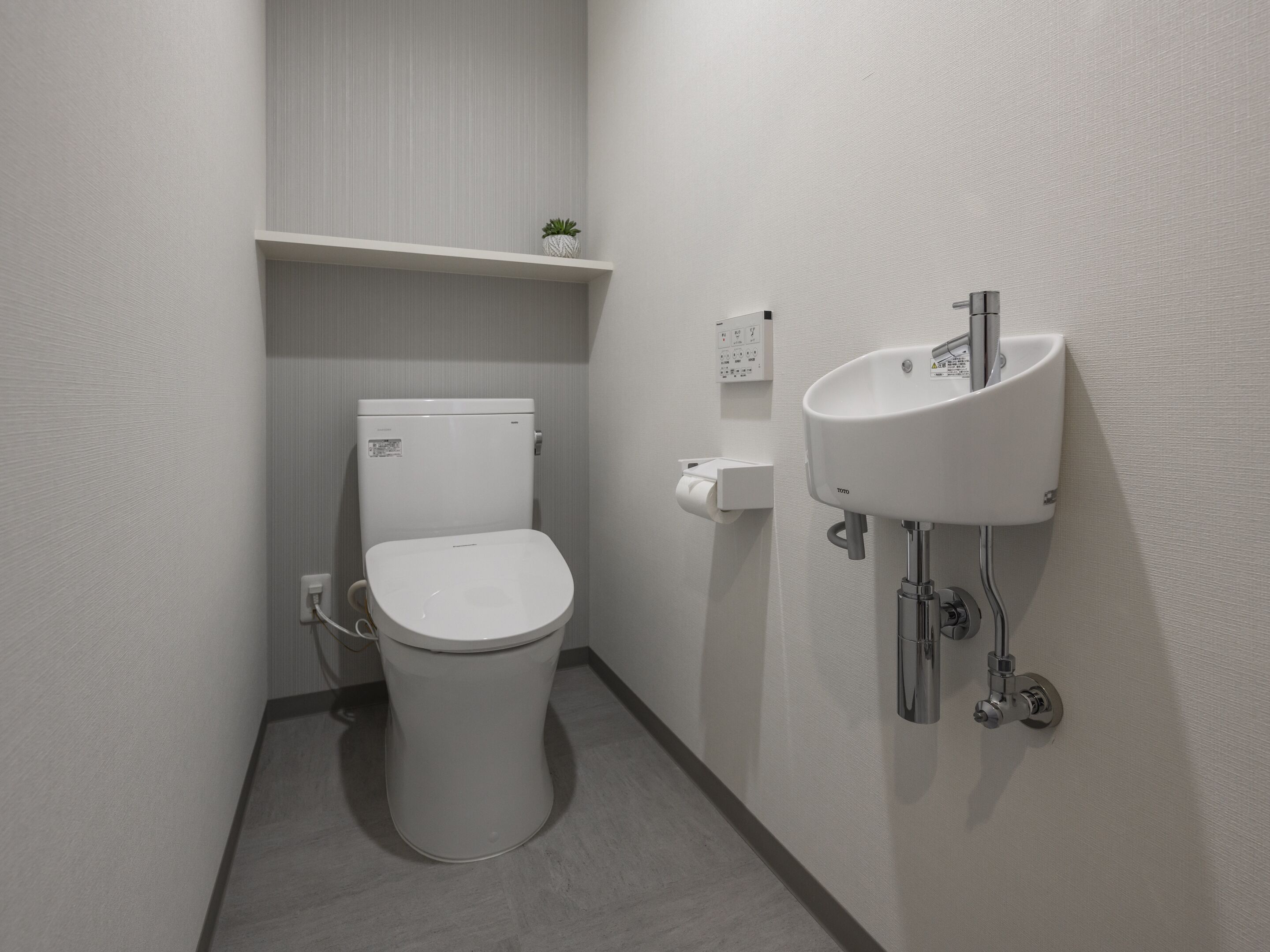 Une salle de bain moderne et compacte, équipée de toilettes, d’un lavabo fixé au mur et d’une petite étagère sur laquelle est posée une plante.