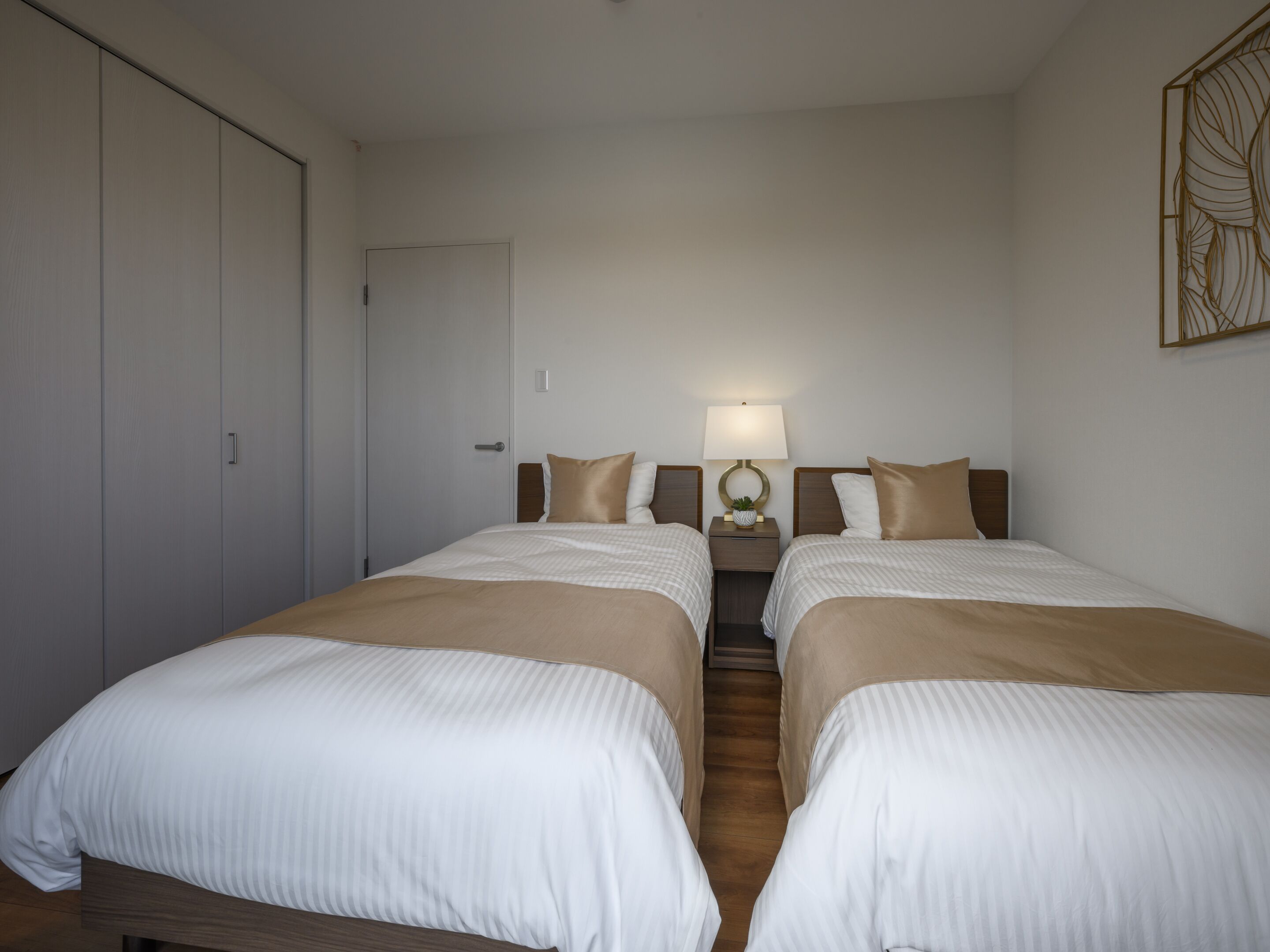 A hotel room with two beds, a wardrobe, a nightstand with a lamp, and a decorative wall piece.