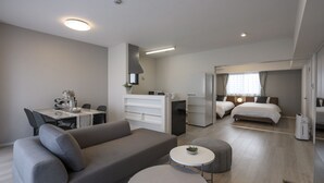 A modern hotel room with a grey sofa, a small round table, a bed with a wooden headboard, a desk with a chair, and a kitchenette area. - &Monde. Okinawa Arena Condominium (Okinawa)