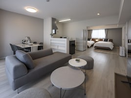 Una habitación de hotel moderna con un sofá gris, una pequeña mesa redonda, una cama con cabecera de madera, un escritorio con silla y una zona de kitchenette.