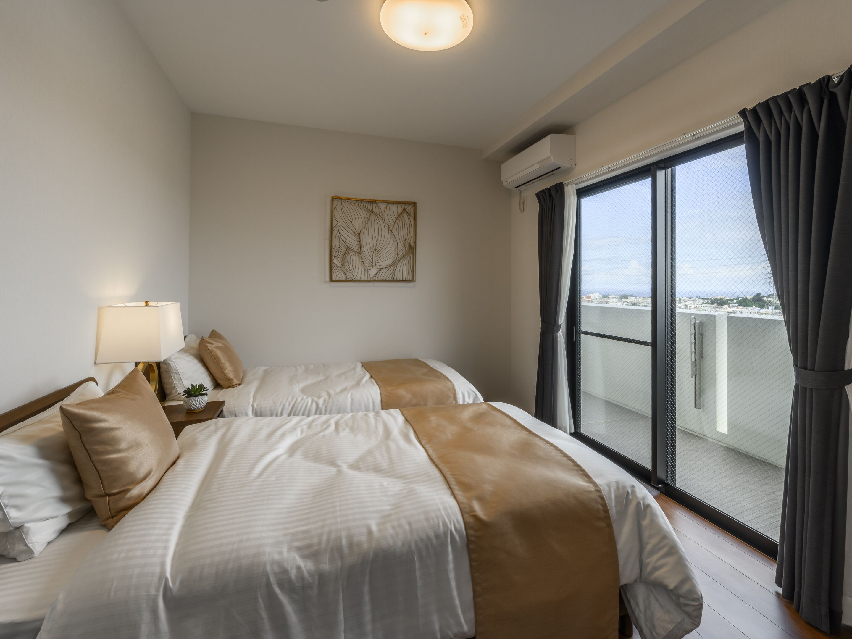 A hotel room with two beds, a bedside table with a lamp, a wall-mounted air conditioner, and a large window with curtains.
