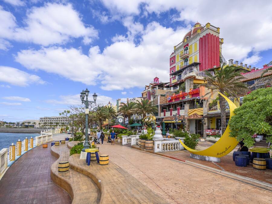 A waterfront promenade with a curved wooden walkway, palm trees, and a variety of shops and restaurants.