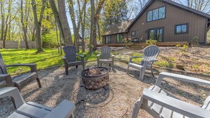 A patio area with Adirondack chairs, a fire pit, and a wooden deck leading to a modern house with large windows. - Belacasa Where Modern Comfort Meets the Quiet of the Woods (Douglas)