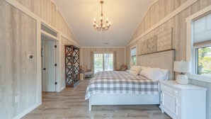 A bedroom with a large bed, a chandelier, a white dresser, and a view of the outdoors. - Belacasa Where Modern Comfort Meets the Quiet of the Woods (Douglas)