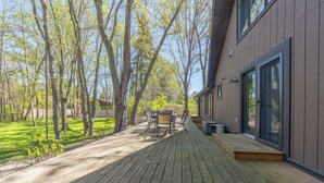 Une terrasse en bois avec une table et des chaises, entourée d’arbres et d’un bâtiment doté de grandes fenêtres.