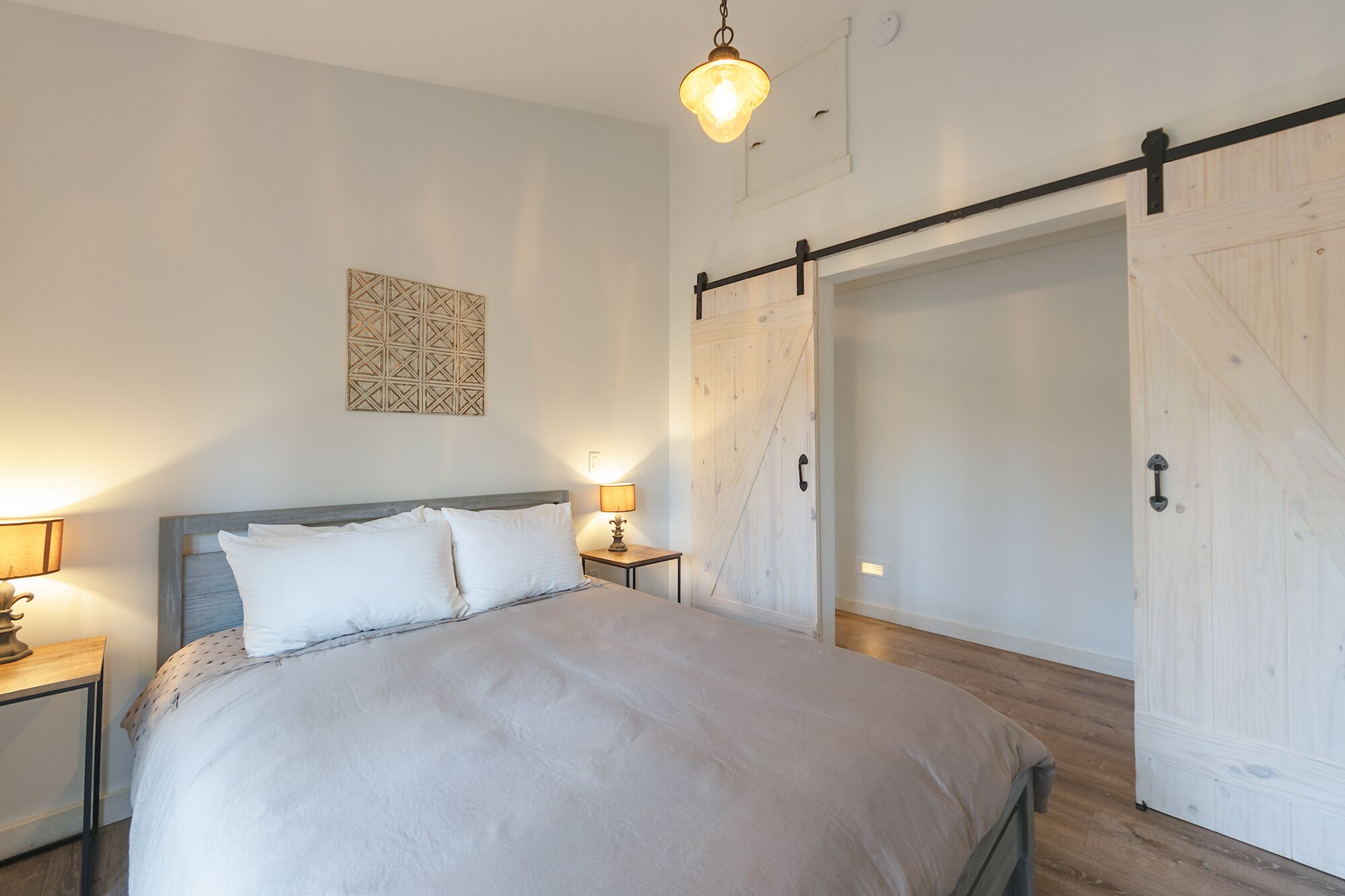 Un dormitorio con cama, mesitas de noche, lámpara colgante, un adorno en la pared y una puerta corredera estilo granero.