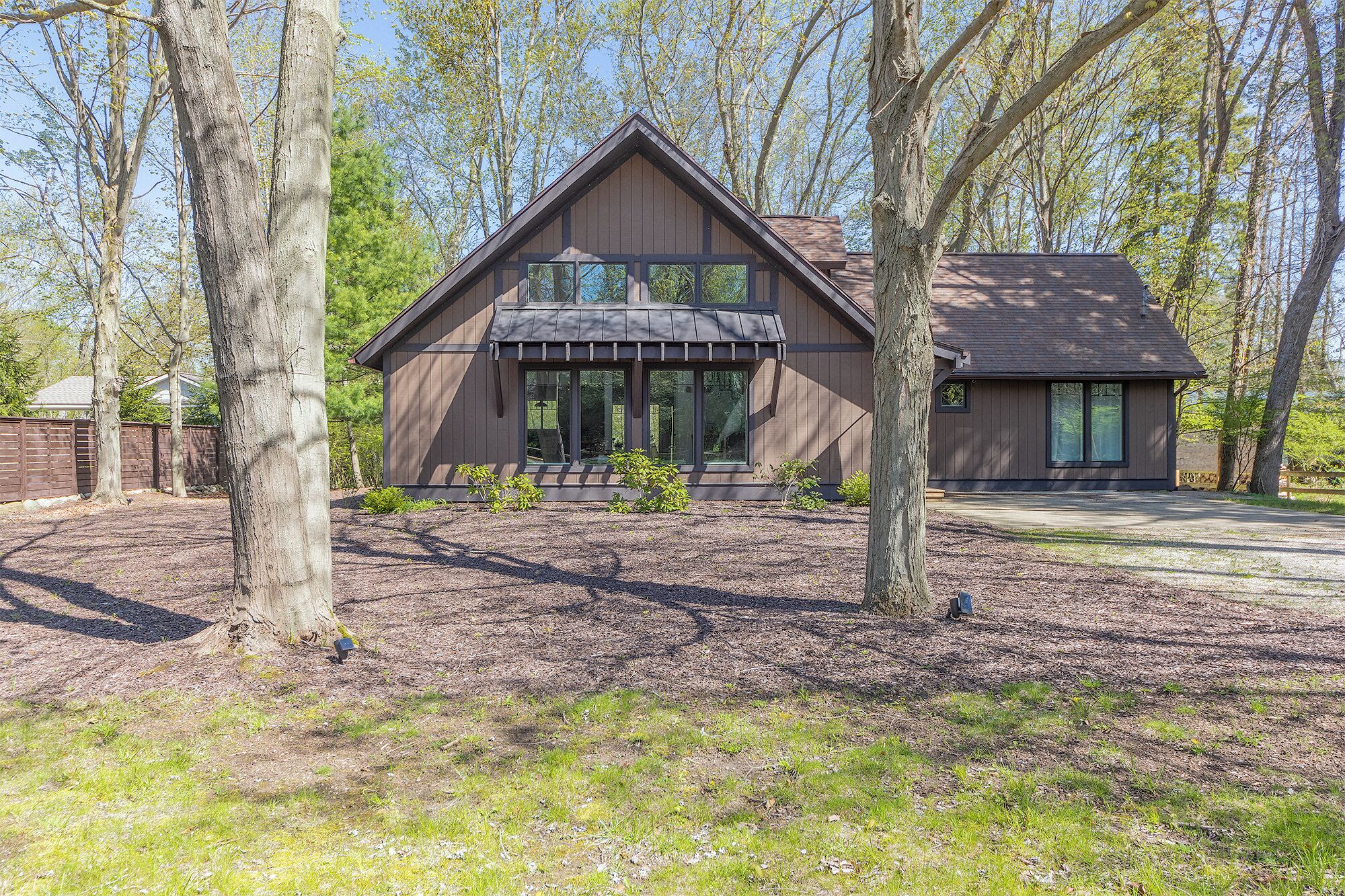 Een modern huis met twee verdiepingen, een ruime veranda, omringd door bomen en een verzorgd gazon.