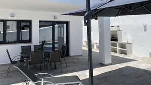 Um pátio moderno ao ar livre com mesa e cadeiras, um guarda-sol grande e vista para uma paisagem montanhosa.