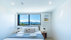 Et moderne soveværelse med en stor seng, to sengeborde, et vindue med udsigt til et bjerglandskab og et billede i ramme på væggen.