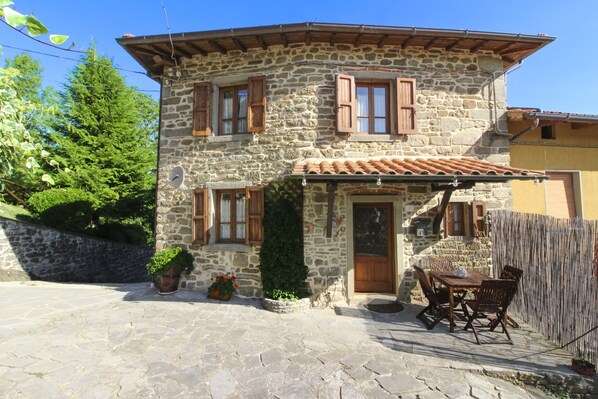 A stone house with a tiled roof, wooden shutters, and a patio with a table and chairs.