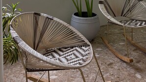Une pièce avec deux fauteuils à bascule en osier, une plante en pot et une plante suspendue près d’une fenêtre donnant sur des arbres et des bâtiments.