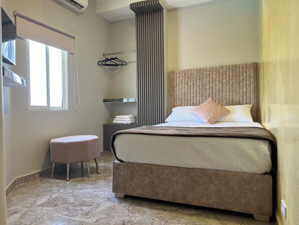 A modern bedroom with a large bed, a small stool, a towel rack, and a window.