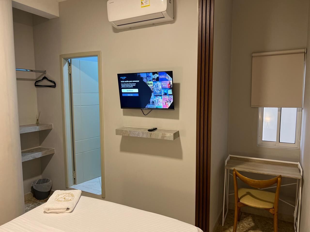 Een hotelkamer met een bed, een aan de muur bevestigde televisie, een klein tafeltje met een stoel en een raam met een rolgordijn.