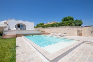 A swimming pool with lounge chairs, a stone wall, and a modern building with an arched entrance.