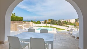 An outdoor patio with a pool, white furniture, and a view of the sea and buildings.