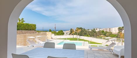 Une terrasse extérieure avec une piscine, des meubles blancs et une vue sur la mer et les bâtiments.
