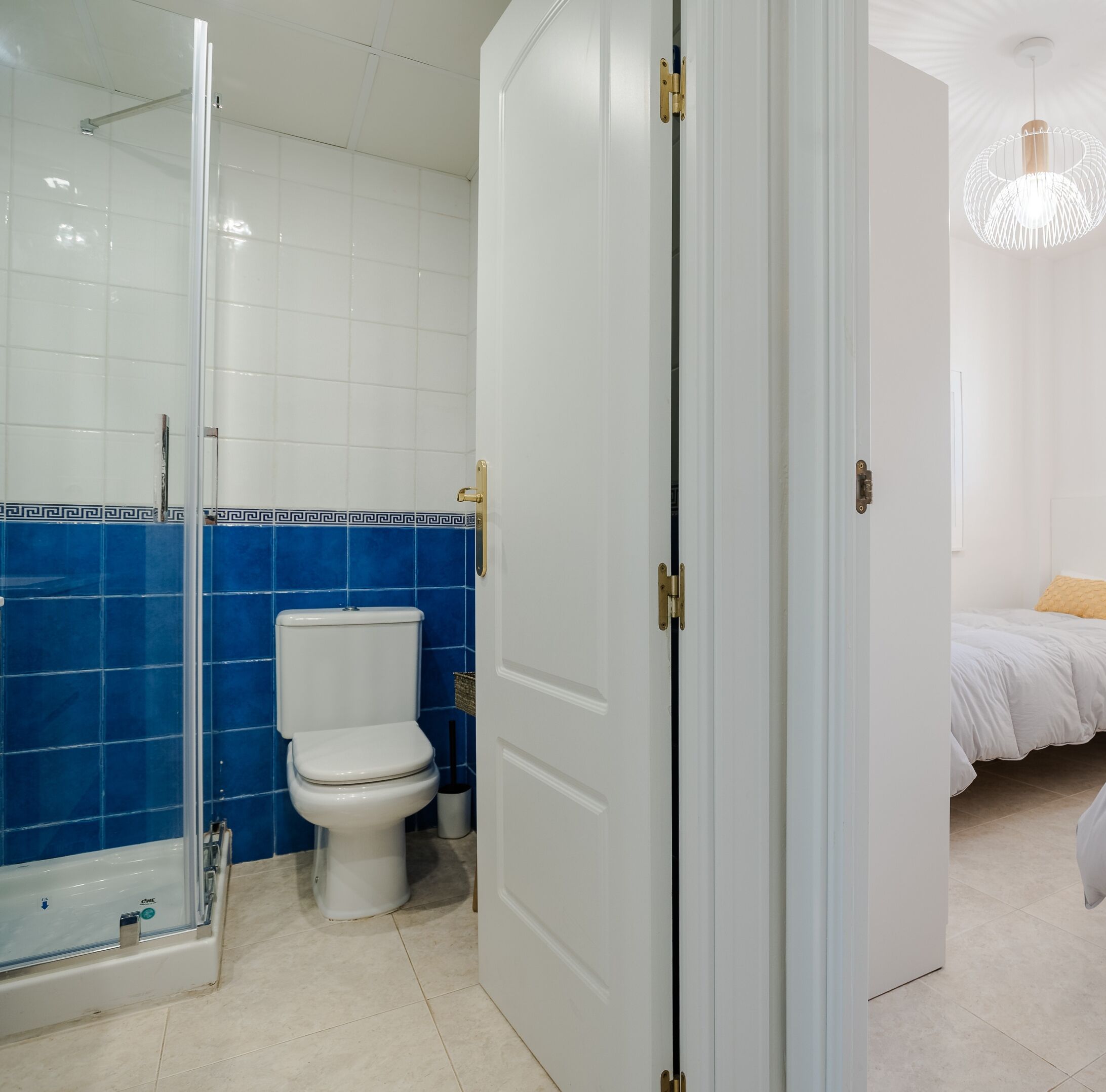 A bathroom with a glass shower enclosure, a toilet, and a bed in the adjacent room.