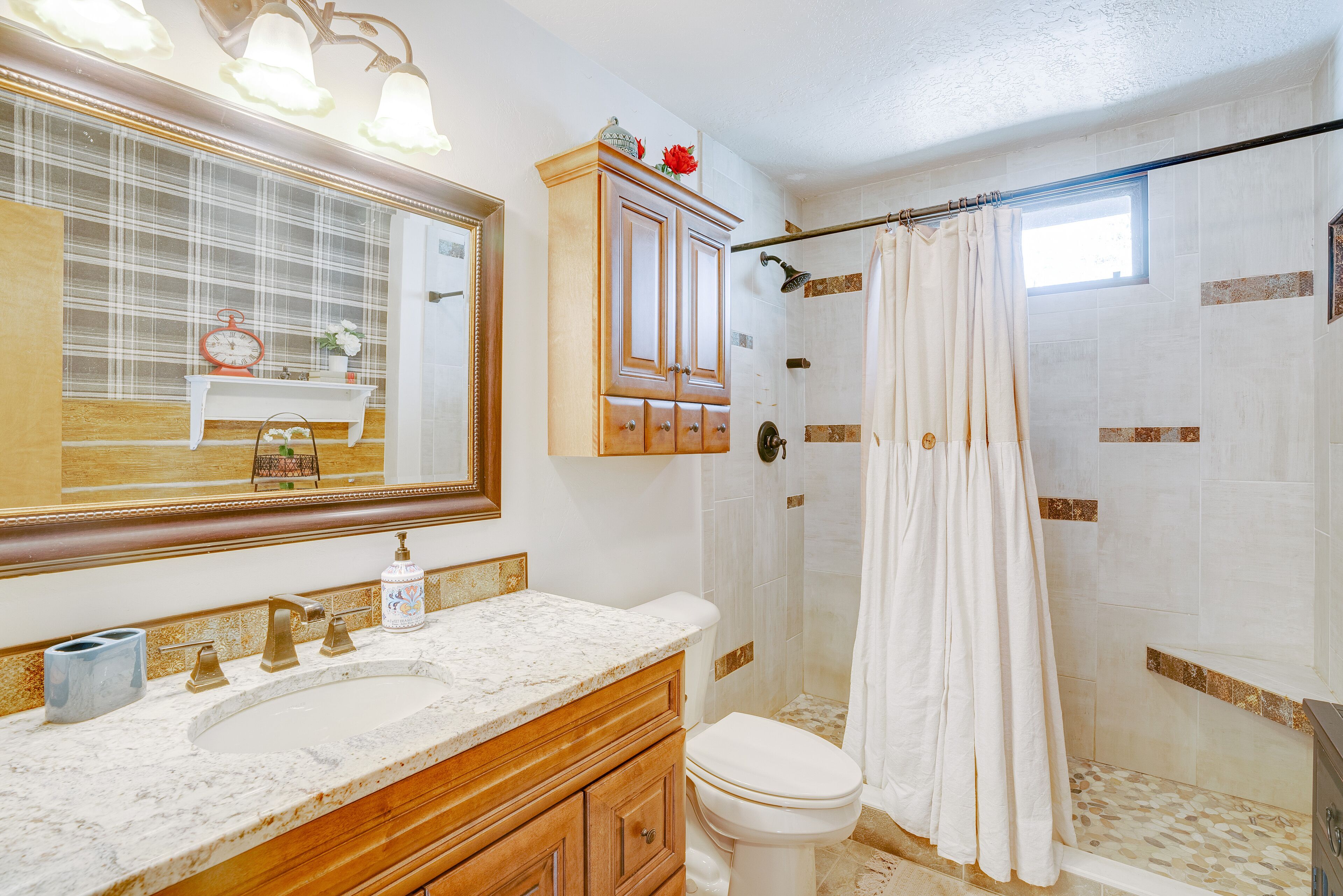A bathroom with a marble sink, wooden vanity, a large mirror, a shower with a curtain, and a window.