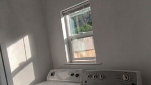 A compact laundry room with a front-loading washer and dryer, a window with blinds, and a view of greenery outside. - Charming Palo Alto Home Next to Stanford (Palo Alto)