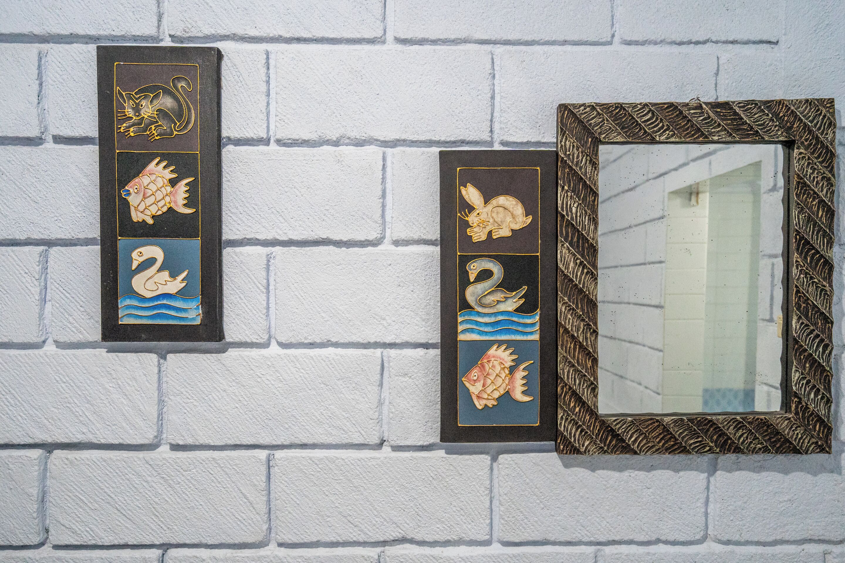 Two decorative plaques with animal designs and a mirror with a patterned frame on a white brick wall.