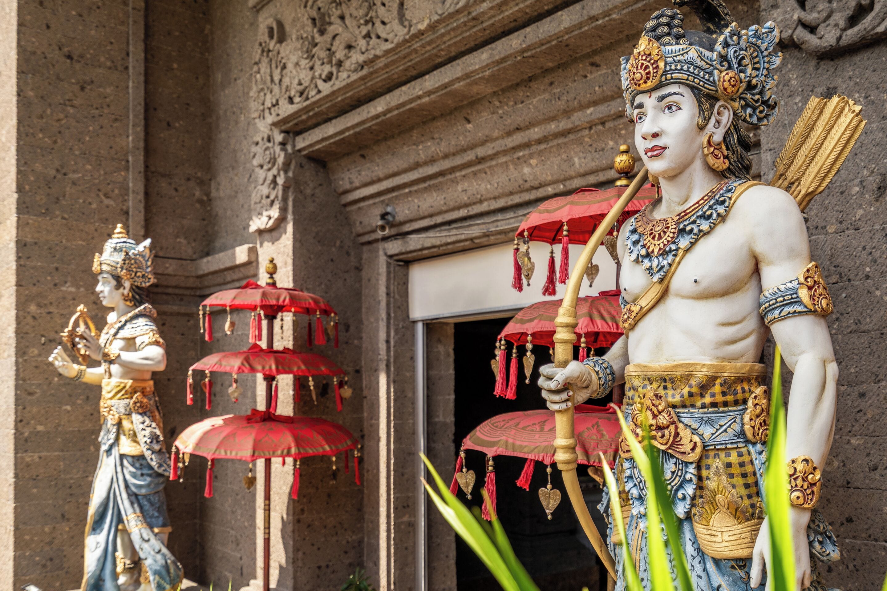A statue of a figure holding a bow and arrow, standing in front of a building with ornate carvings and red umbrellas.