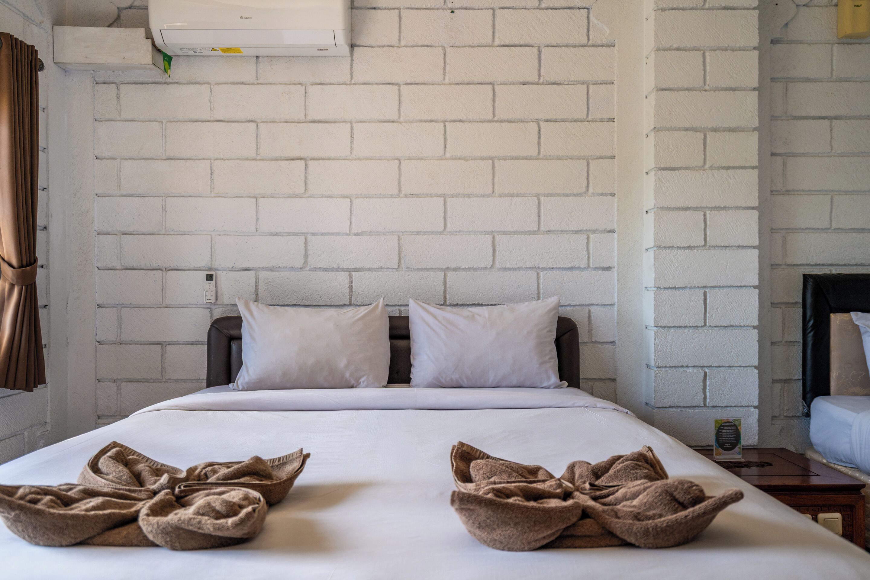 A hotel room with a brick-patterned wall, a bed with white linens, two pillows, and a bedside table with a lamp.