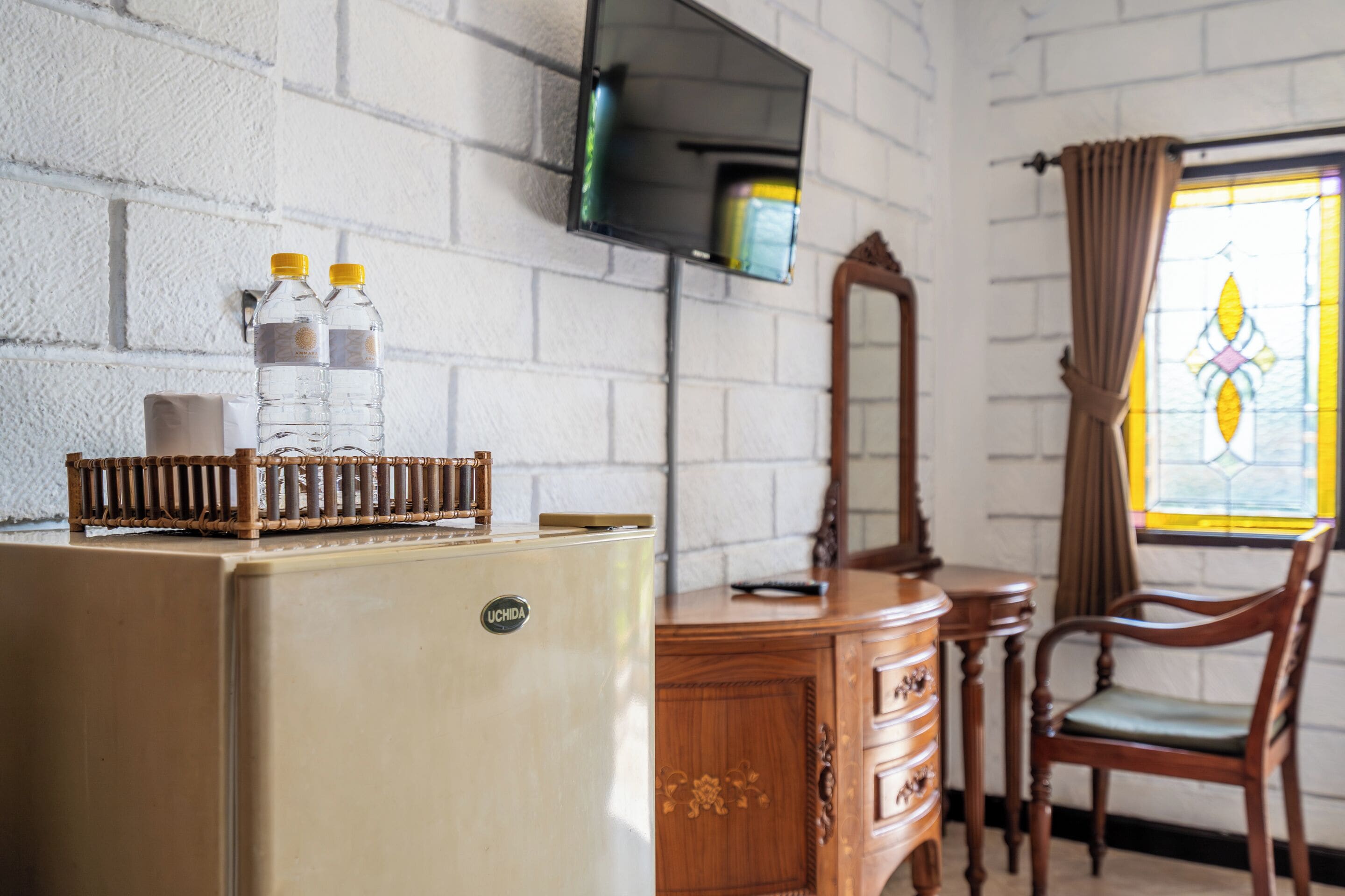A room with a white brick wall, a wooden dresser, a chair, a television mounted on the wall, and a window with curtains.