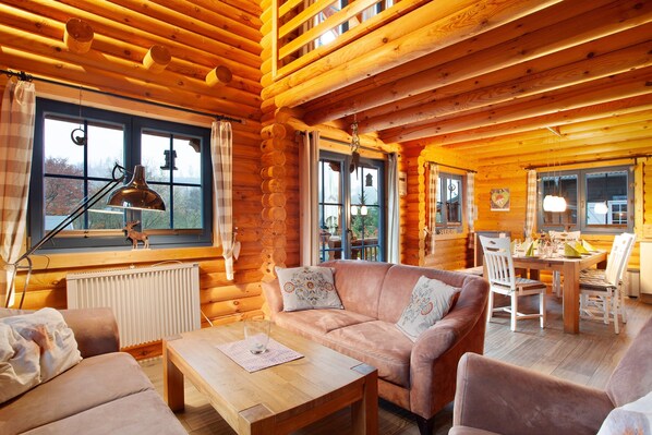 A log cabin interior with a wooden floor, a sofa, a dining table, and a window with a view.