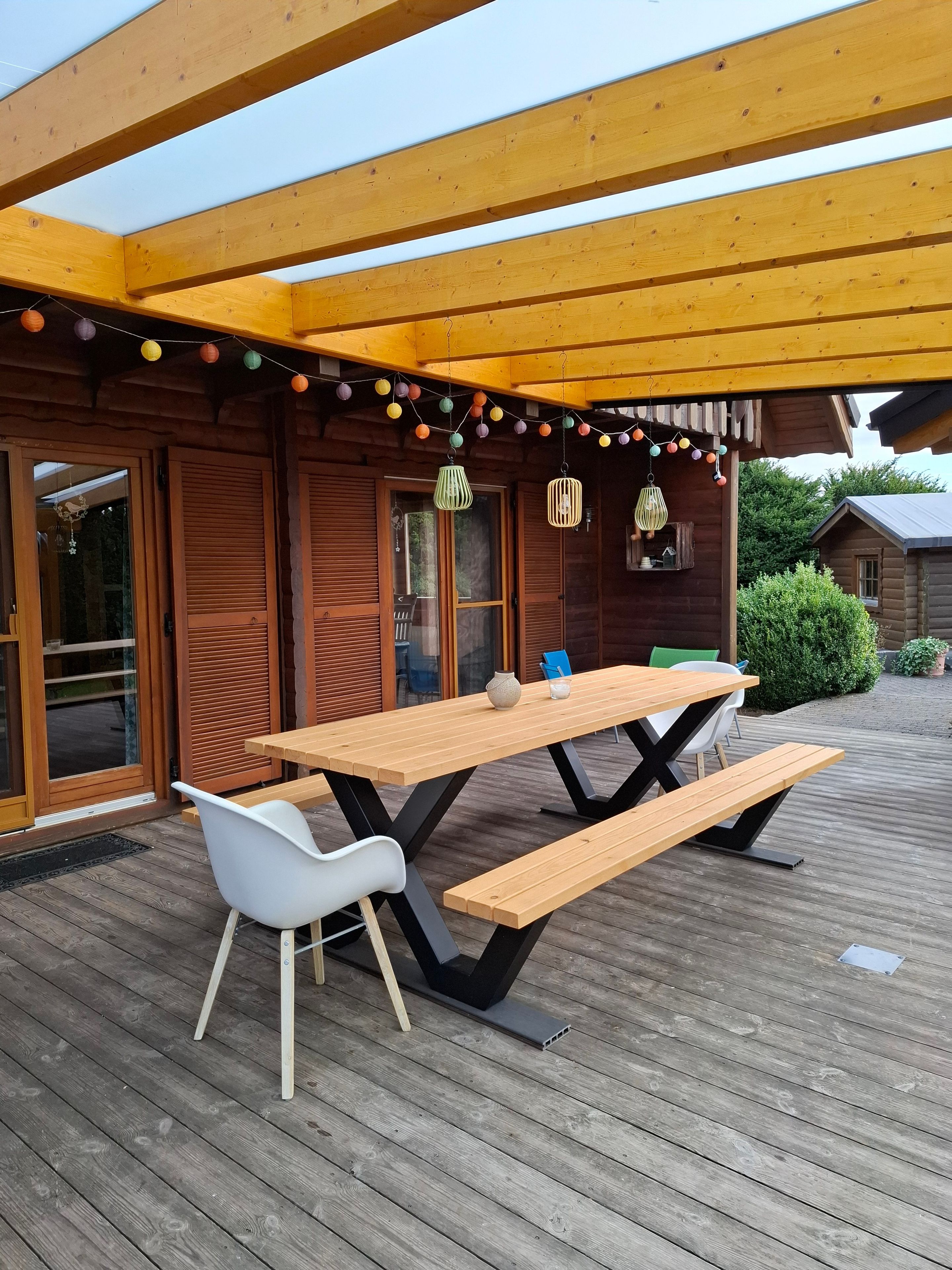 En utomhusveranda av trä med ett picknickbord och stolar, omgiven av träväggar och ett takförsett område med ljusslingor.