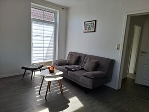 Un salon avec un canapé gris, une petite table blanche et une fenêtre avec des stores.