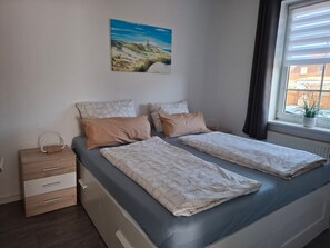 Une chambre avec deux lits, une table de chevet avec une lampe, une fenêtre avec des rideaux et un tableau au mur.