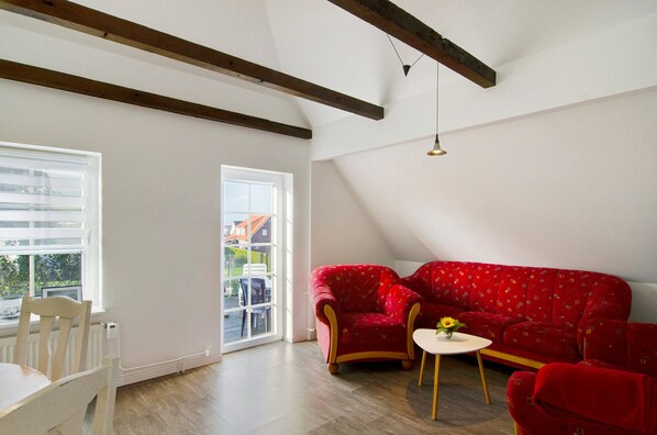 Una habitación con un sofá rojo estampado, una mesita con un florero, una silla blanca y una ventana con persianas.