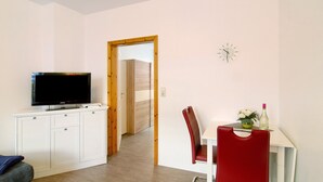 A room with a television on a white cabinet, a dining table with a red chair, and a clock on the wall.