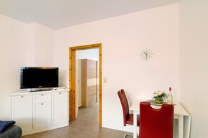 A room with a television on a white cabinet, a dining table with a red chair, and a clock on the wall.
