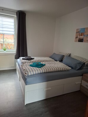 Un dormitorio con una cama, una ventana con cortinas, un radiador, una pared con un cuadro y una planta pequeña en el alféizar.