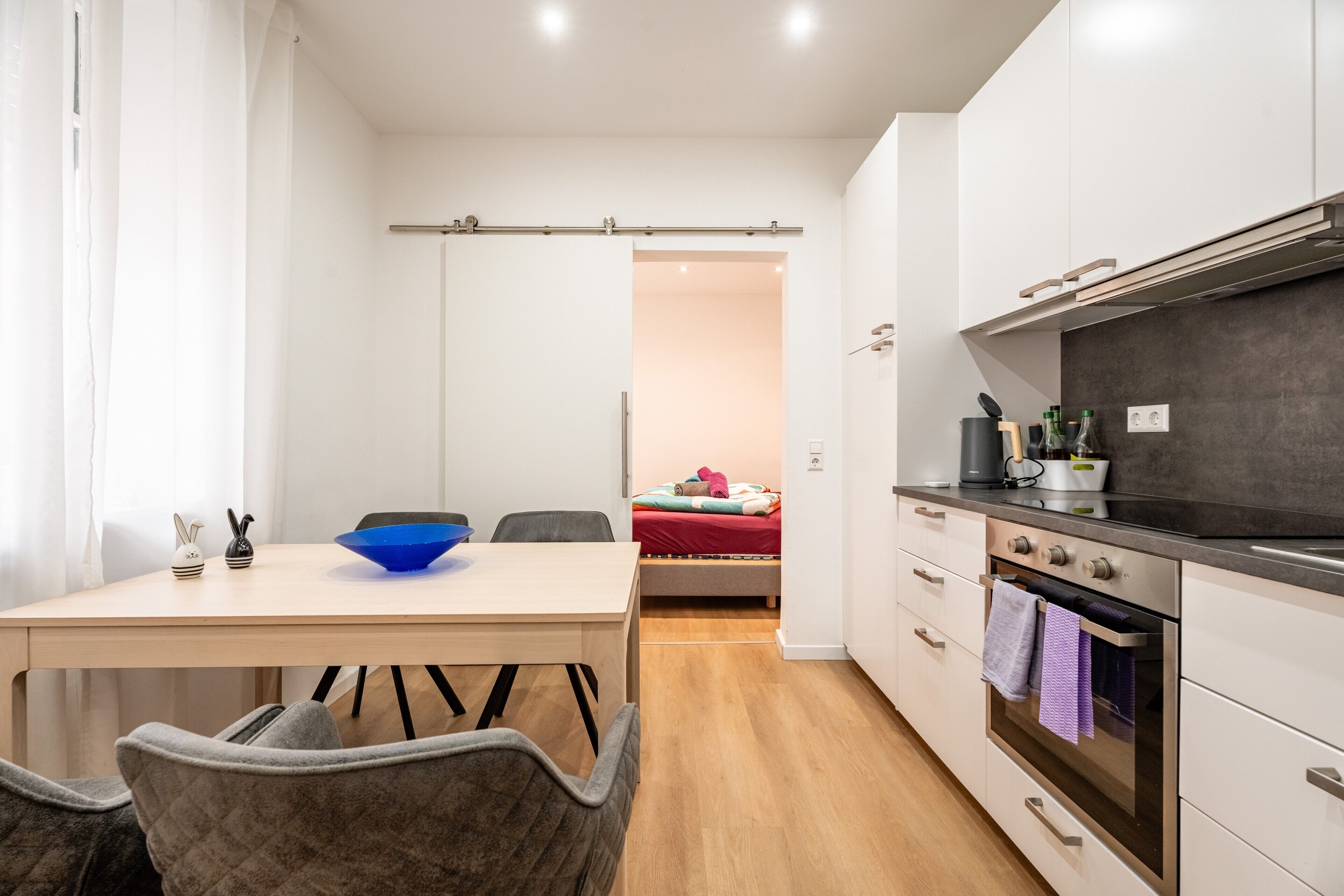 Cocina moderna con comedor, una cama al fondo y un divisor corredero que da a otra habitación.
