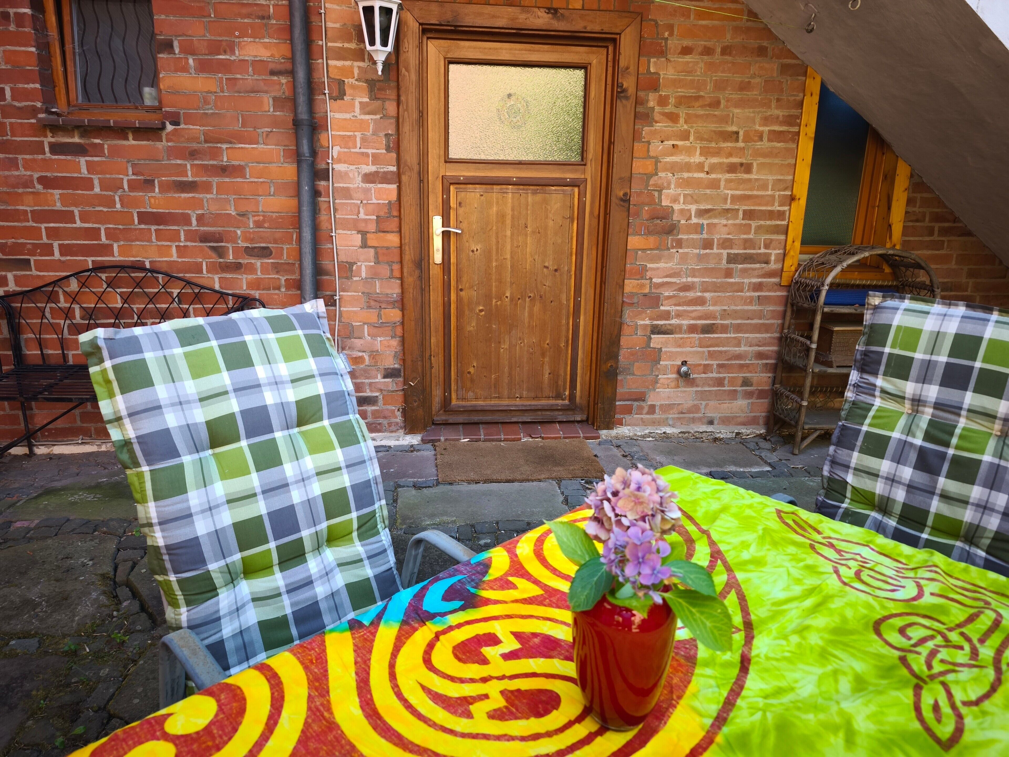 Un edificio de ladrillo con una puerta de madera, dos sillas de metal con cojines a cuadros y una mesa con mantel colorido y un jarrón con flores.