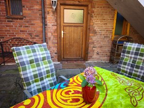 A brick building with a wooden door, two metal chairs with plaid cushions, and a table with a colorful tablecloth and a vase with flowers.